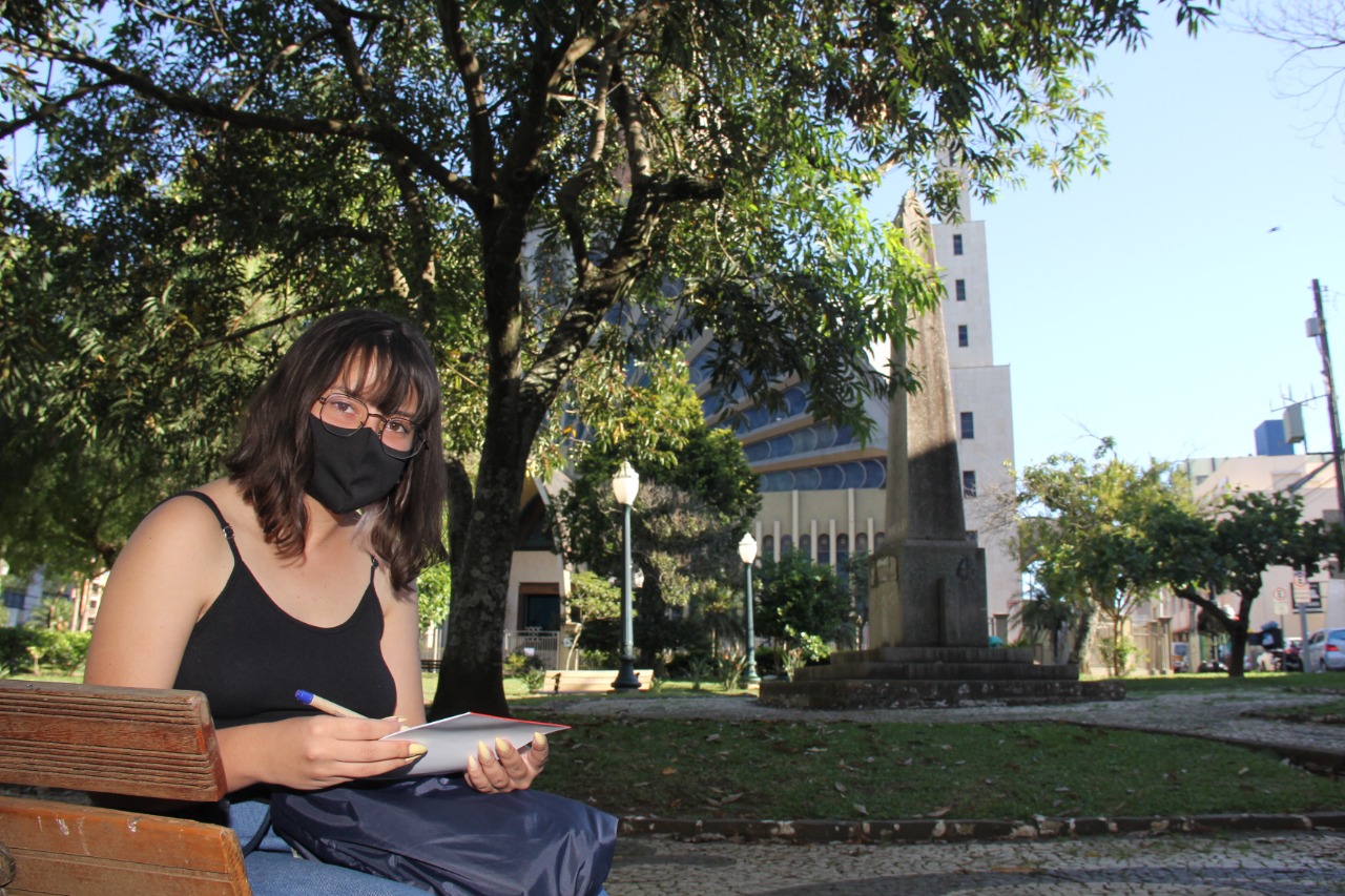 Foto de Érica Gabriele Boiano, sentada em banco da Praça Marechal Floriano Peixo, tendo ao fundo o obelisco que homenageia os fundadores de Ponta Grossa