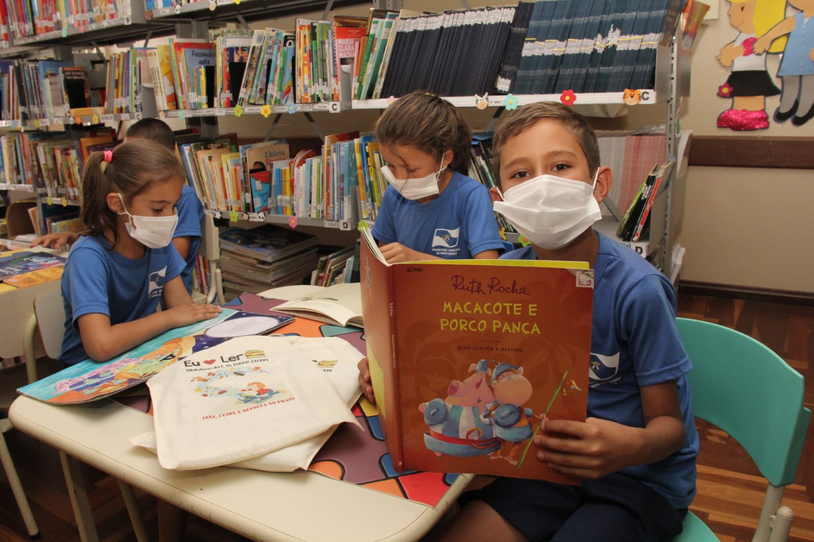 Foto de David de Souza Dias, aluno de escola de Ponta Grossa, segurando o livro "Macacote e Porco Pança", de Ruth Rocha, dentro de biblioteca, ao lado de suas colegas