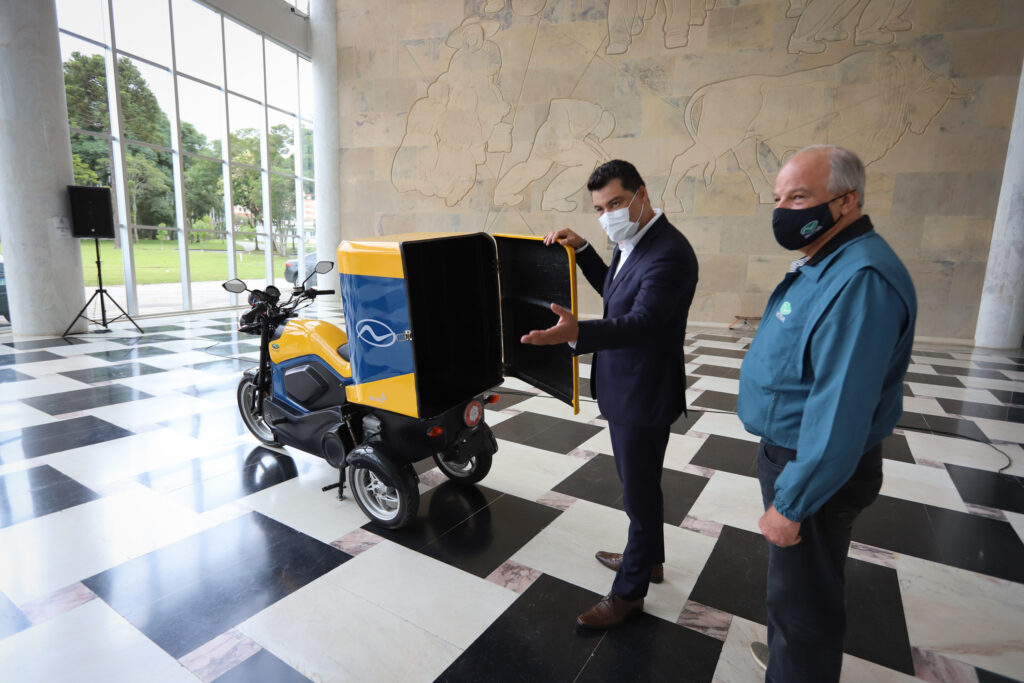 Foto de triciclo híbrido 100% nacional estacionado nas dependências do Palácio do Iguaçu, com Marcelo Rangel e representante da Motiva Motocicletas