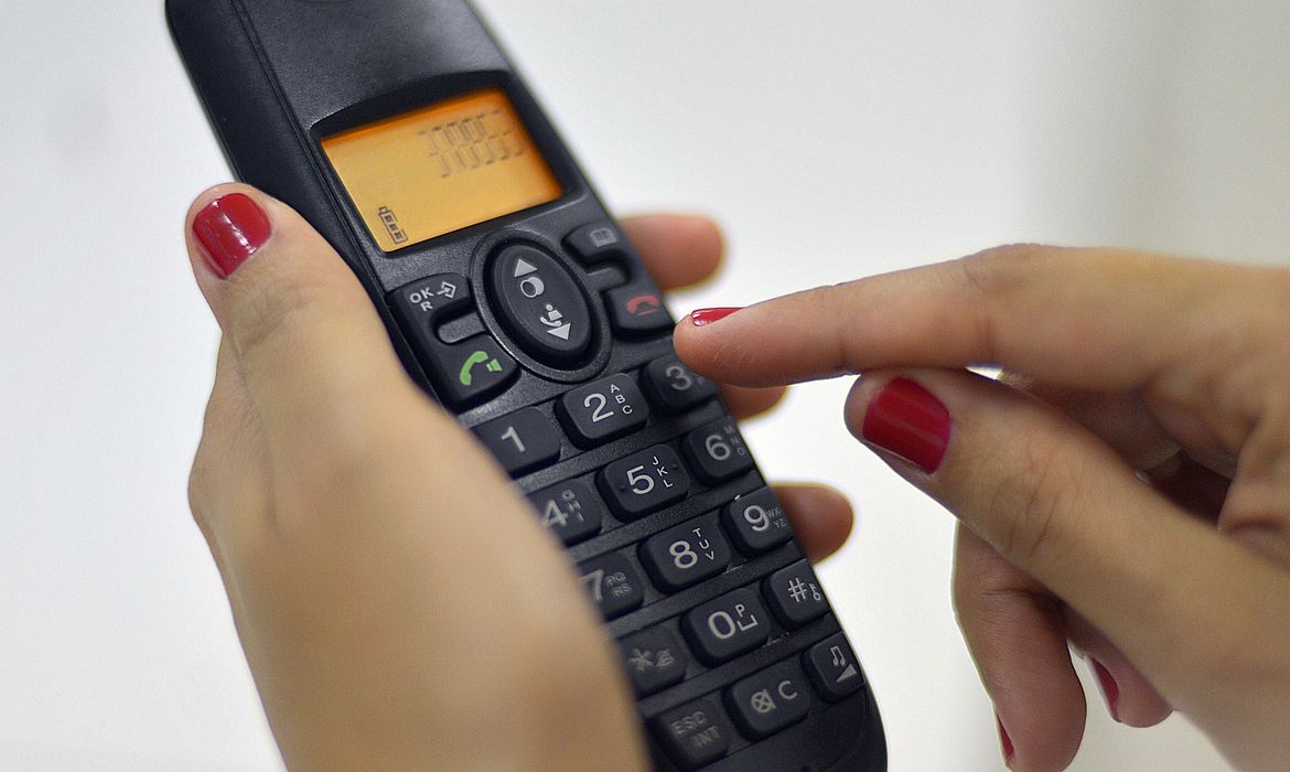 Pessoa discando em um telefone fixo com um número na tela.