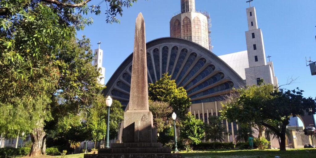 Foto da Praça Marechal Floriano Peixoto, em Ponta Grossa, mostrando obelisco em homenagem a fundadores e Catedral Sant'Ana