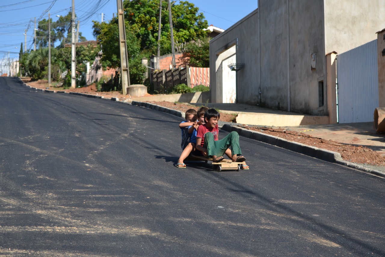 Três crianças descendo em cima de um carrinho de rolimã em rua inclinada