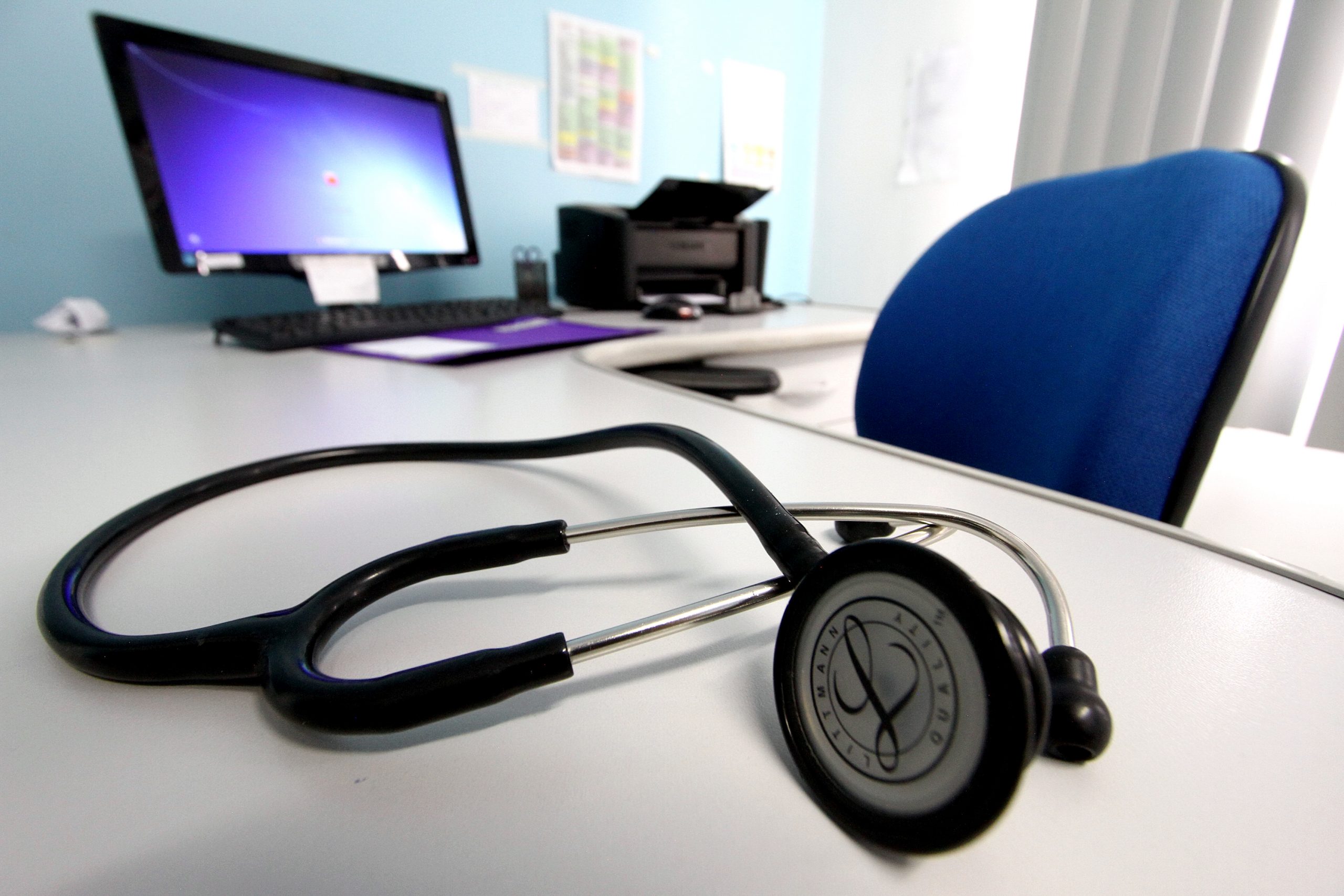 Estetoscópio sobre a mesa de um médico, tendo ao fundo um computador e uma impressora