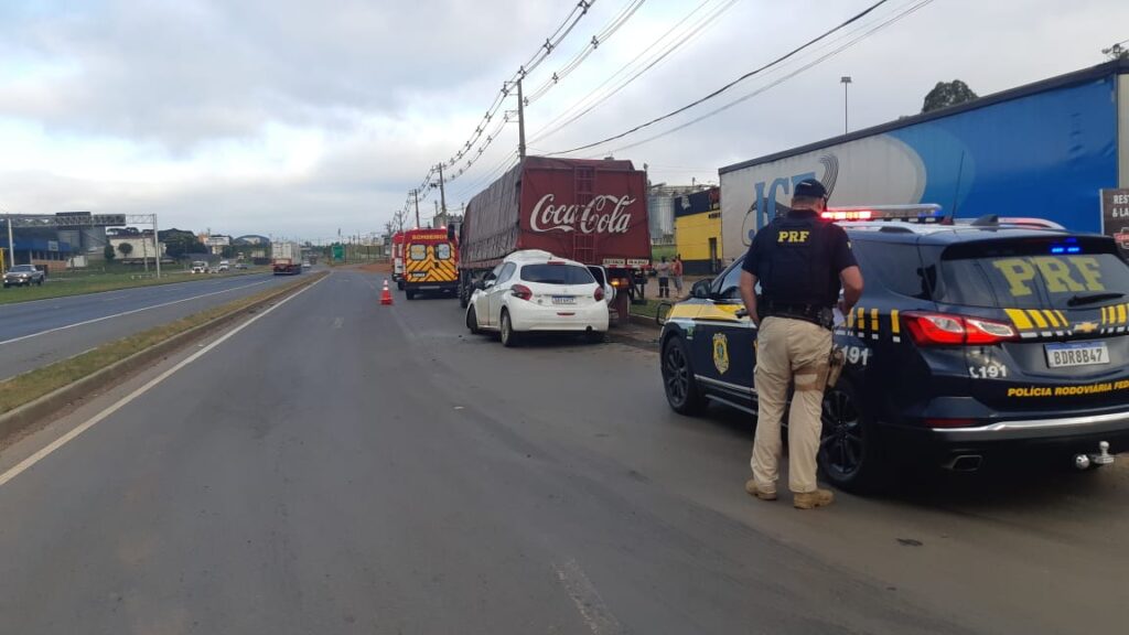 Foto de policial rodoviário federal, ao lado de viatura, em local de acidente envolvendo Peugeot 208 e caminhão da Coca-Cola