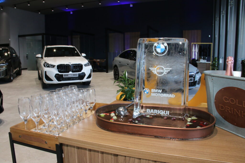Placa de vidro da BMW em um balcão, ao lado de taças e, ao fundo, veículos branco e prata da BMW