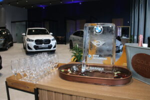 Placa de vidro da BMW em um balcão, ao lado de taças e, ao fundo, veículos branco e prata da BMW