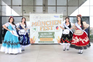 Rainha e princesas da Münchenfest posam diante do banner do evento.