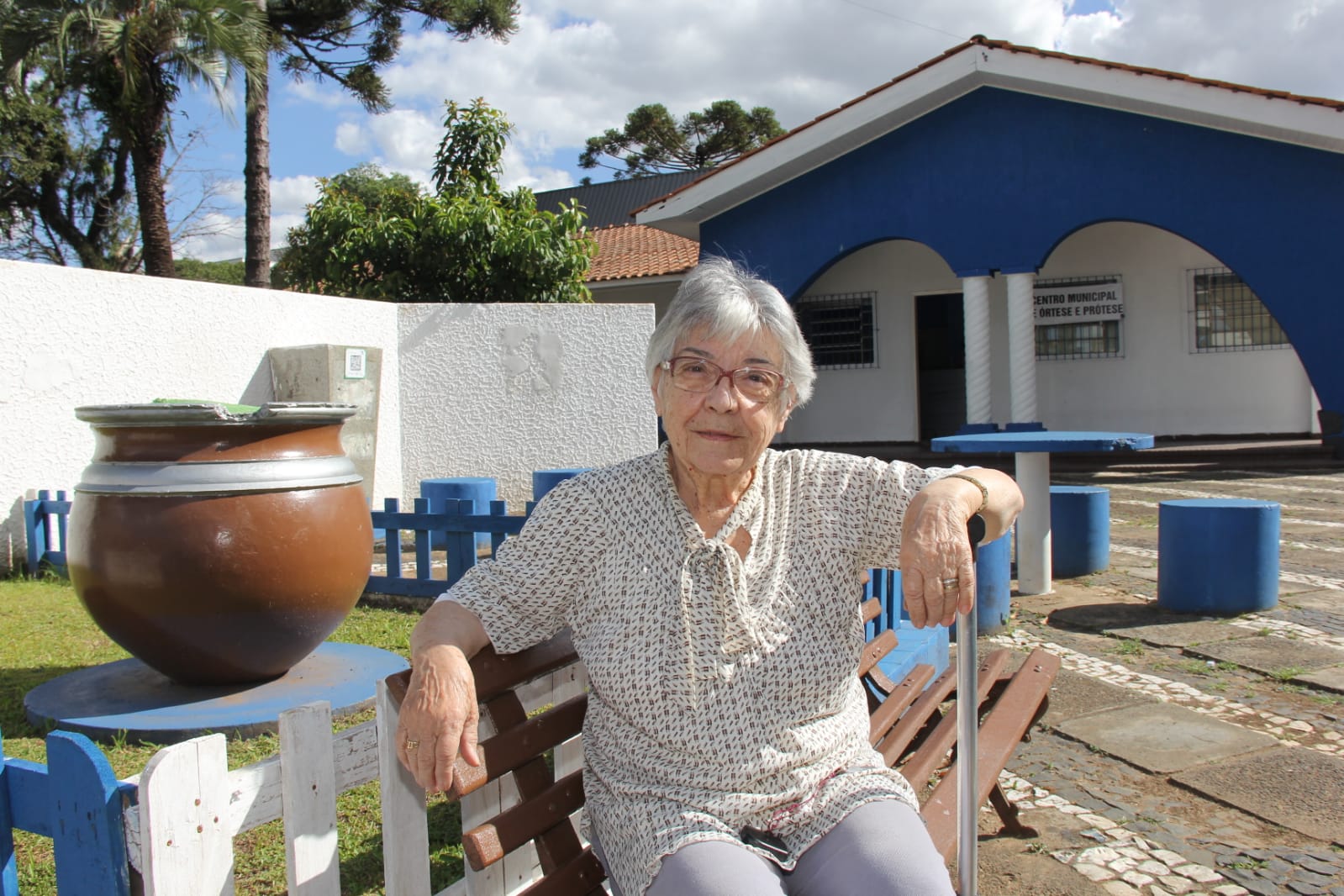 Foto de Isaura Carvalho Pauluk, sentada em banco da Praça Getúlio Vargas, ao fundo estrutura mais antiga desde a criação da primeira praça no local