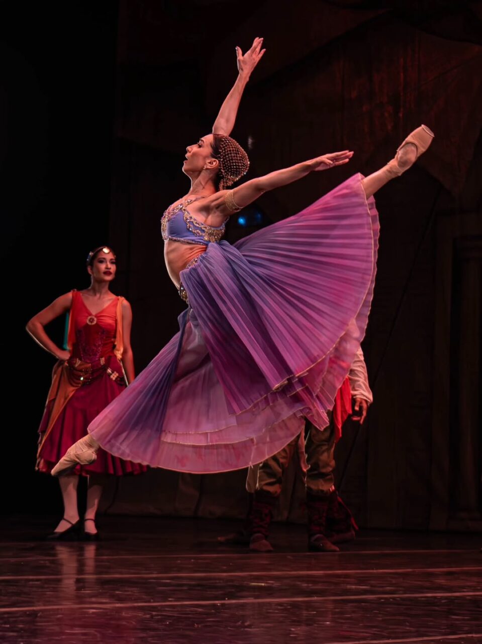 Bailarina Juliana Valadão em passo de balé no ar vestida com figurino colorido.