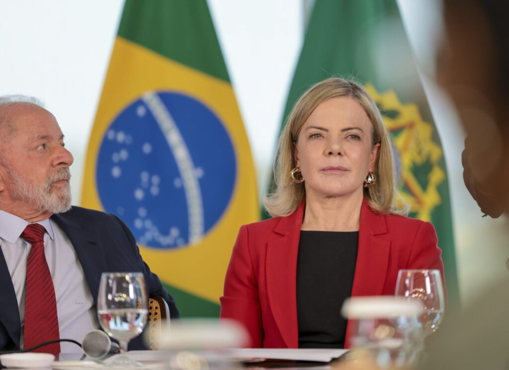 Presidente Lula e ministra Gleisi sentados, com a bandeira do Brasil ao fundo