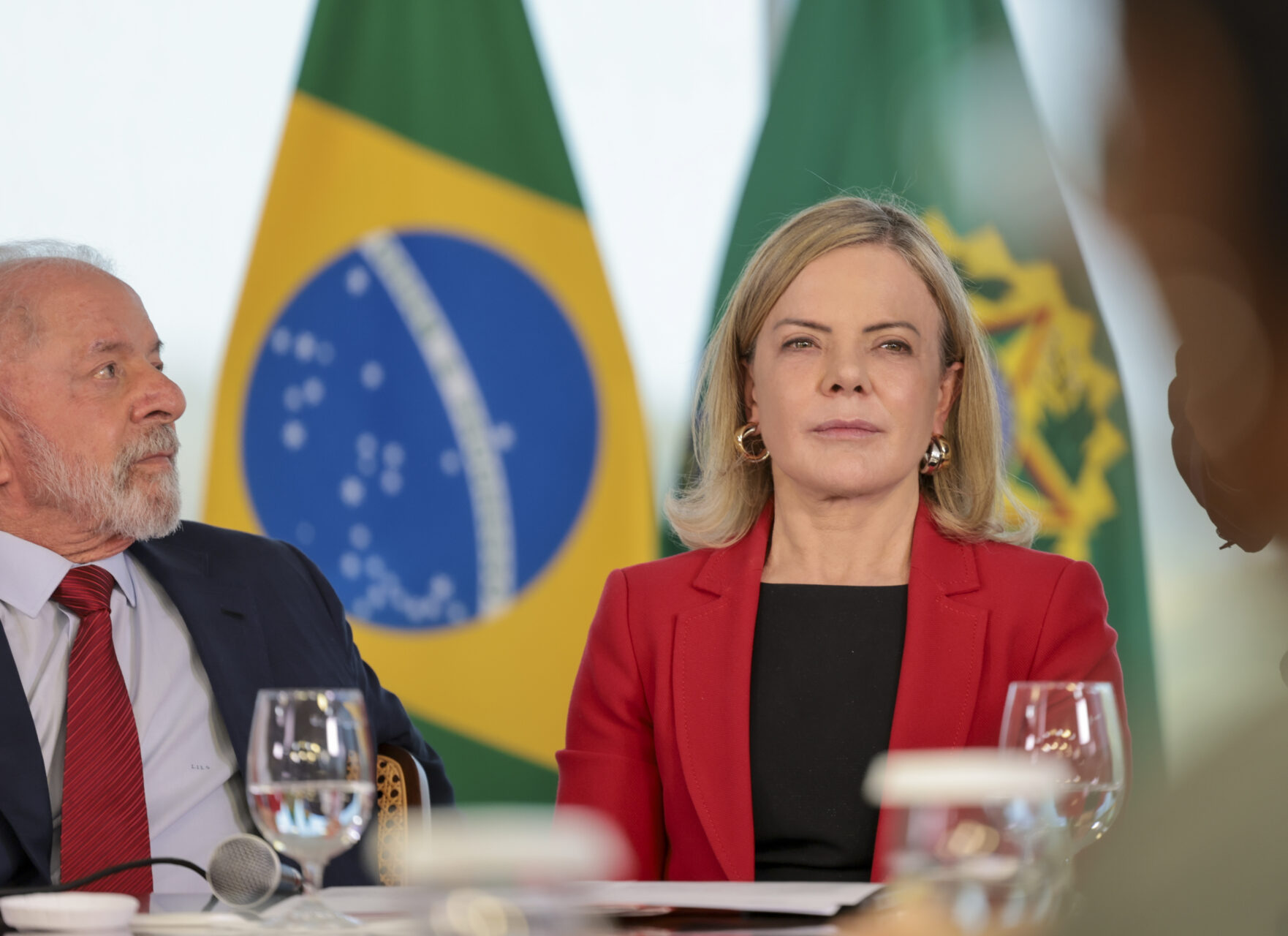Presidente Lula e ministra Gleisi sentados, com a bandeira do Brasil ao fundo