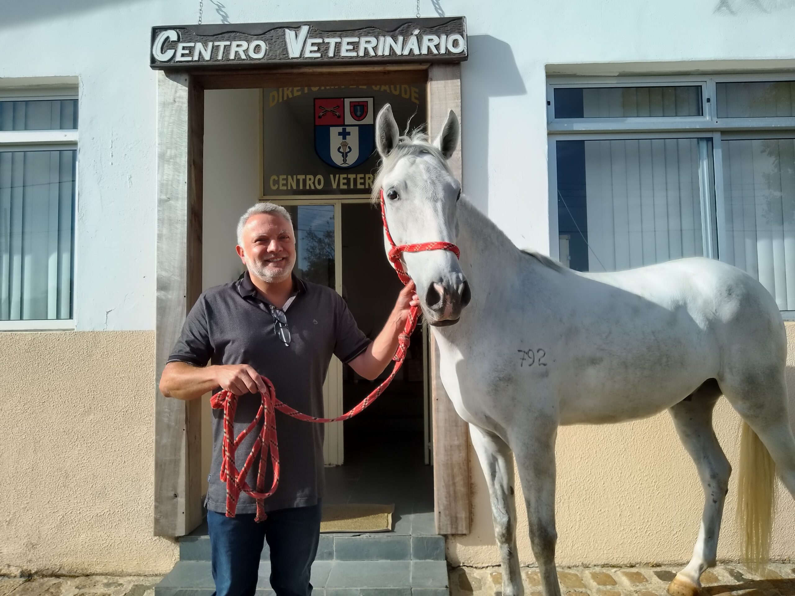 Médico veterinário posa para foto com um cavalo branco. E atrás está o Centro Veterinário do Regimento.
