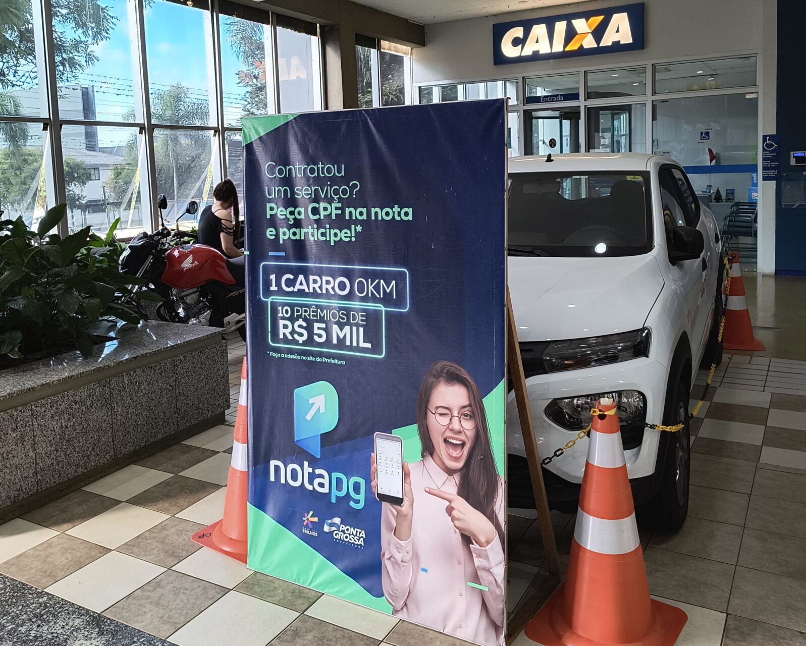 Automóvel que será sorteado no IPTU Premiado de Ponta Grossa, parcialmente oculto atrás de banner da campanha