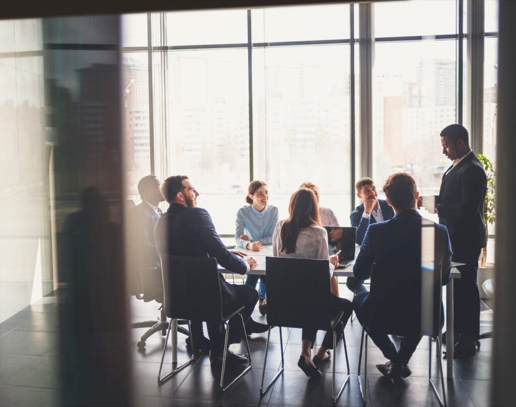 Grupo de pessoas participando de uma reunião diante e uma mesa, em ambiente corporativo, com grandes janelas de vidro que mostram edifícios ao fundo