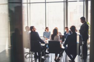 Grupo de pessoas participando de uma reunião diante e uma mesa, em ambiente corporativo, com grandes janelas de vidro que mostram edifícios ao fundo