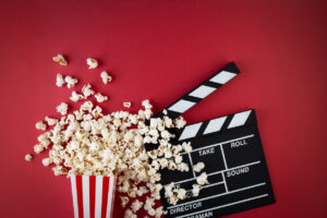 Foto ilustrativa da Semana do Cinema tem pacote de pipocas e claquete em fundo vermelho.