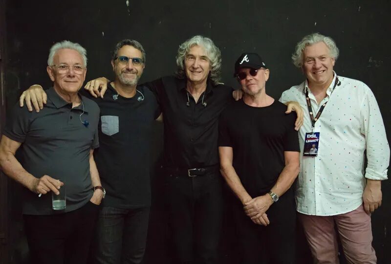 Membros da banda Dire Straits Legacy posam para foto em fundo preto.