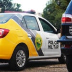 Foto ilustrativa mostrando uma viatura da Polícia Militar do Paraná, em uma estrada com árvores e céu acinzentado ao fundo