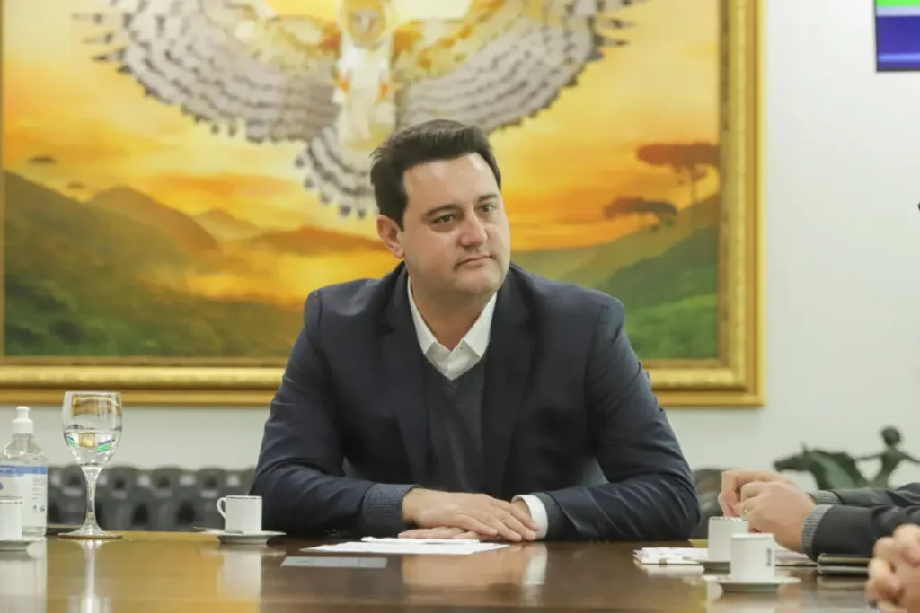 Governador Ratinho Junior, sentado, com os braços sobre a mesa, olhando para a direita com atenção
