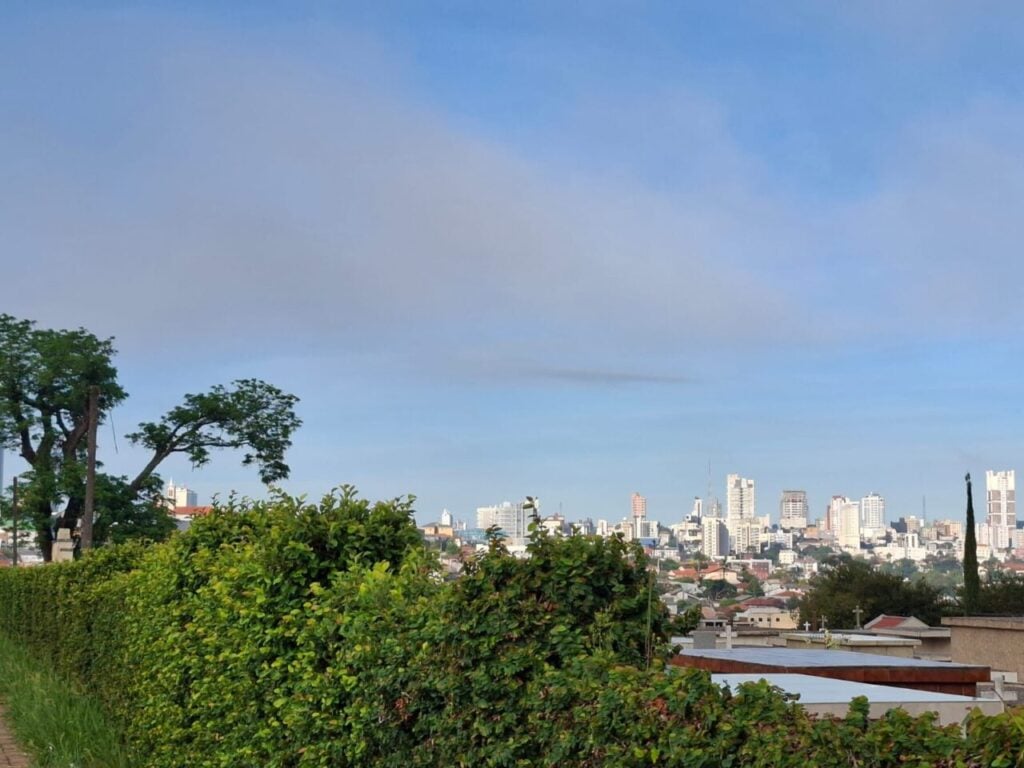 Céu parcialmente nublado em Ponta Grossa, com árvores em primeiro plano e prédios da cidade ao fundo ilustram previsão do tempo