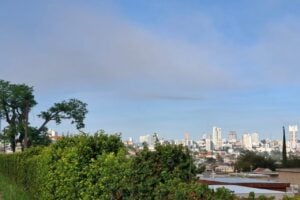 Céu parcialmente nublado em Ponta Grossa, com árvores em primeiro plano e prédios da cidade ao fundo ilustram previsão do tempo