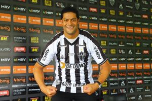 Jogador do Atlético Mineiro posa sorrindo com camisa listrada preta e branca diante de painel de patrocinadores, fazendo pose de força