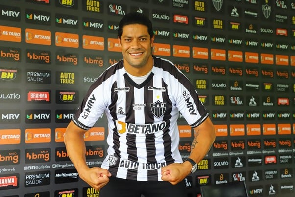 Jogador do Atlético Mineiro posa sorrindo com camisa listrada preta e branca diante de painel de patrocinadores, fazendo pose de força