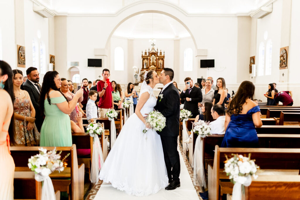 Recém-casados se beijam no interior de uma igreja, cercados por convidados que admiram a cena e fazem o registro com seus celulares