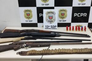 armas de foto sobre uma mesa da delegacia, tendo ao fundo a logomarca da Polícia Civil do Paraná e da 13ªSDP