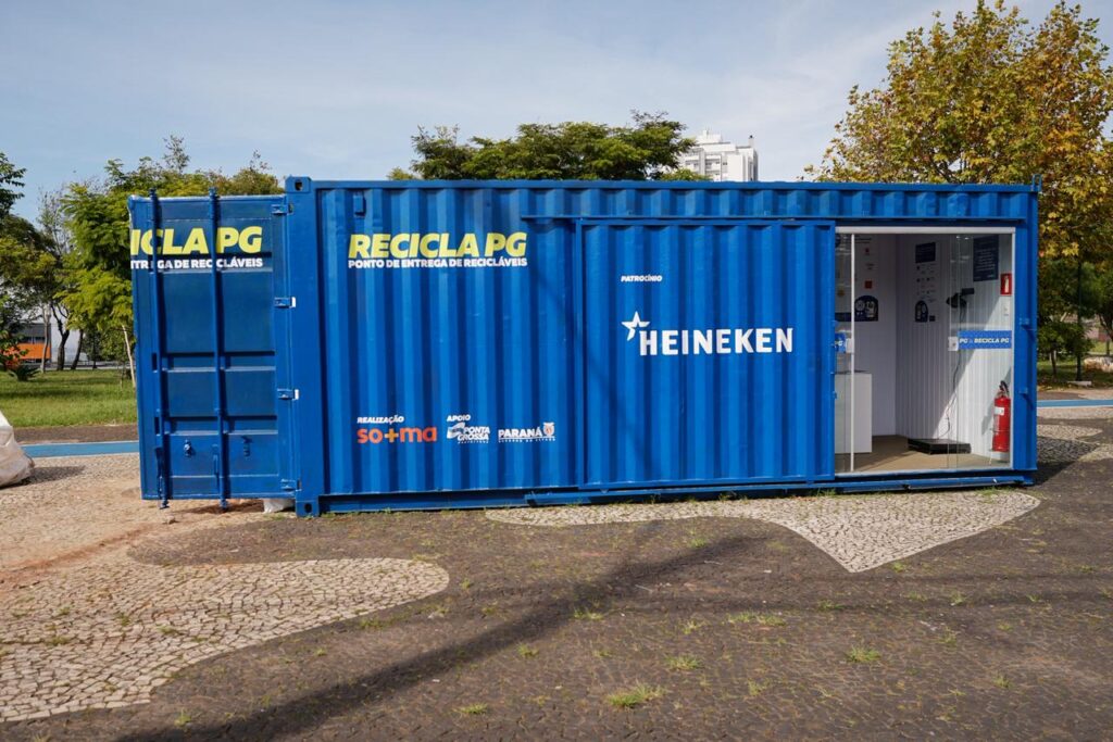 Um contêiner azul adaptado que funciona como ponto de coleta de recicláveis em um espaço ao ar livre. Ele possui a marca “Recicla PG.