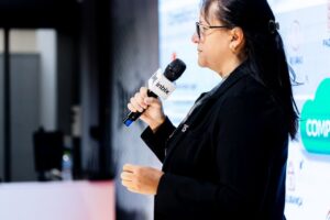Foto de perfil da Advogada Flávia Machado falando em evento, segurando um microfone com sua mão direita