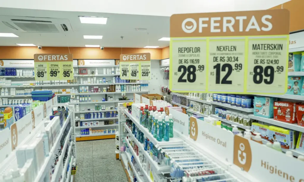 A imagem mostra o interior de uma farmácia bem iluminada e organizada. Há prateleiras cheias de produtos de higiene, cuidados pessoais e medicamentos.