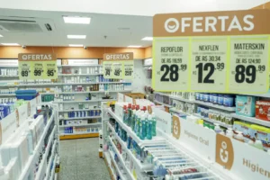 A imagem mostra o interior de uma farmácia bem iluminada e organizada. Há prateleiras cheias de produtos de higiene, cuidados pessoais e medicamentos.