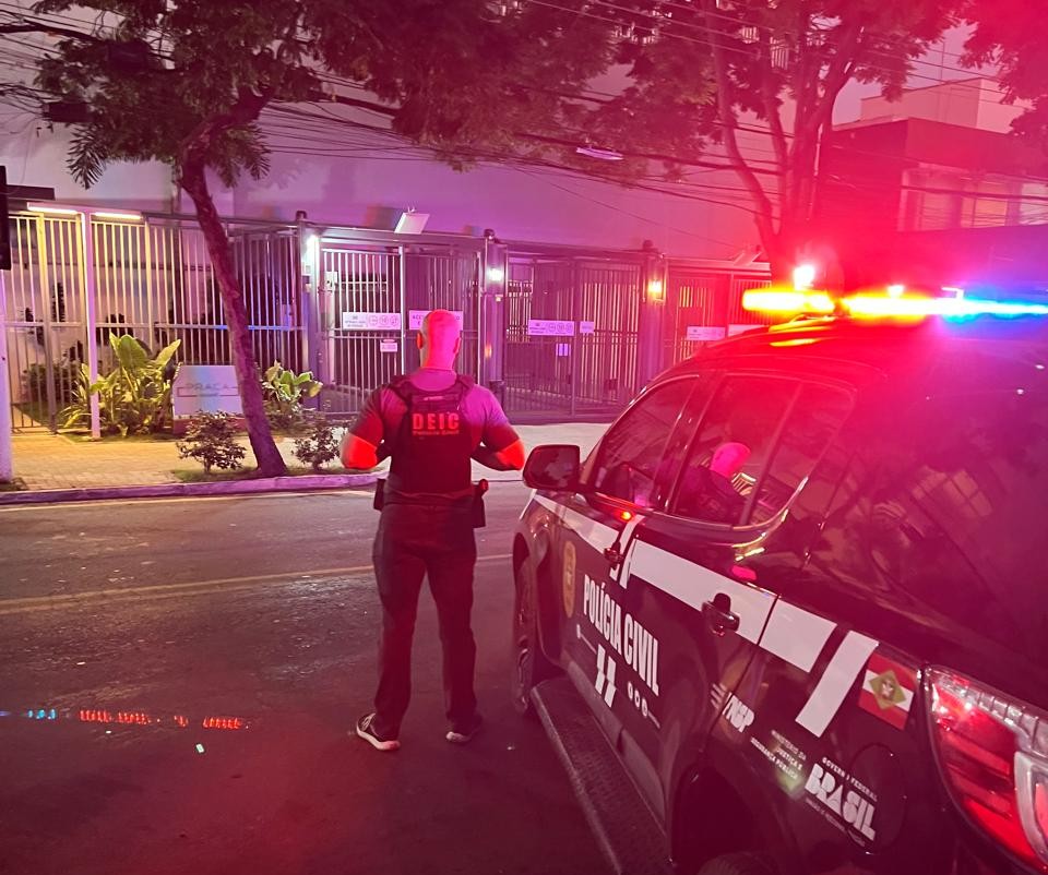 Polícial Civil de costas, ao lado de viatura com o giroflex aceso, em frente à portaria de um prédio, em foto para ilustrar a operação que investiga fraude e lavagem de dinheiro envolveo uso indevido da marca Havan