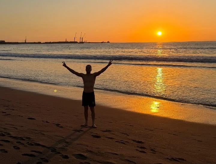 Homem posa para fotografia mostrando o pôr do sol no Oceano Pacífico