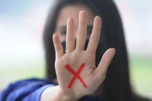 Uma pessoa aparece ao fundo desfocada enquanto estende a mão aberta em direção à câmera. Na palma da mão há um “X” vermelho pintado.