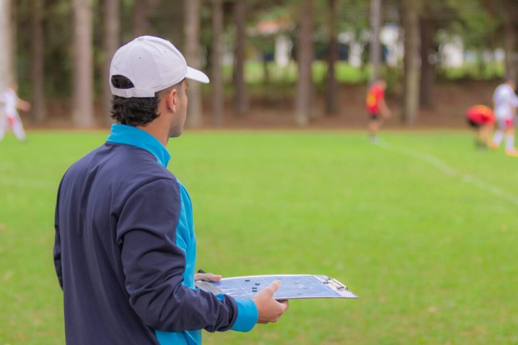Treinador de futebol de costas para a foto, olhando para jogo no gramado e segurando uma prancheta