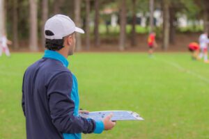 Treinador de futebol de costas para a foto, olhando para jogo no gramado e segurando uma prancheta