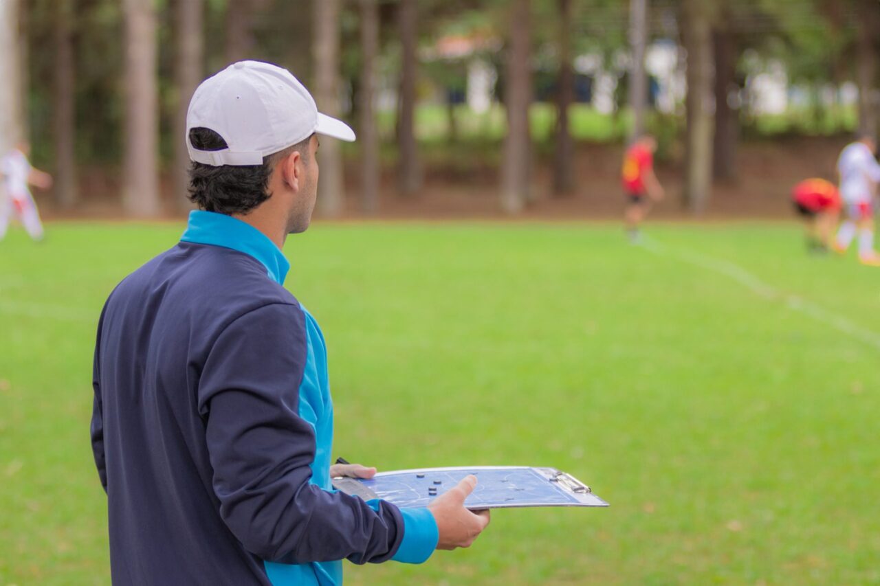 Treinador de futebol de costas para a foto, olhando para jogo no gramado e segurando uma prancheta