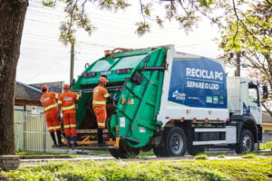 Caminhão de coleta de lixo com três garis se segurando na parte de trás do veículos