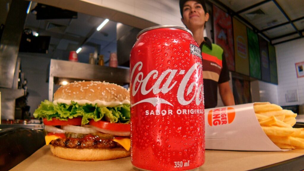 Lata de Coca-Cola em primeiro plano, à frente de um combo de lanches do Burguer King em cima de um balcão, com uma garçonete ao fundo.