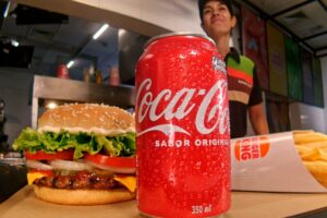 Lata de Coca-Cola em primeiro plano, à frente de um combo de lanches do Burguer King em cima de um balcão, com uma garçonete ao fundo.