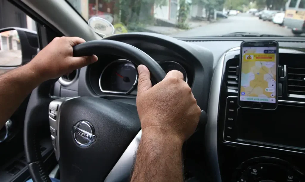 Mãos de motorista de aplicativo em volante de automóvel, tendo fixado ao consolde um smartphone mostrando mapa na tela, sendo um dos possíveis beneficiados pelo programa Move Brasil