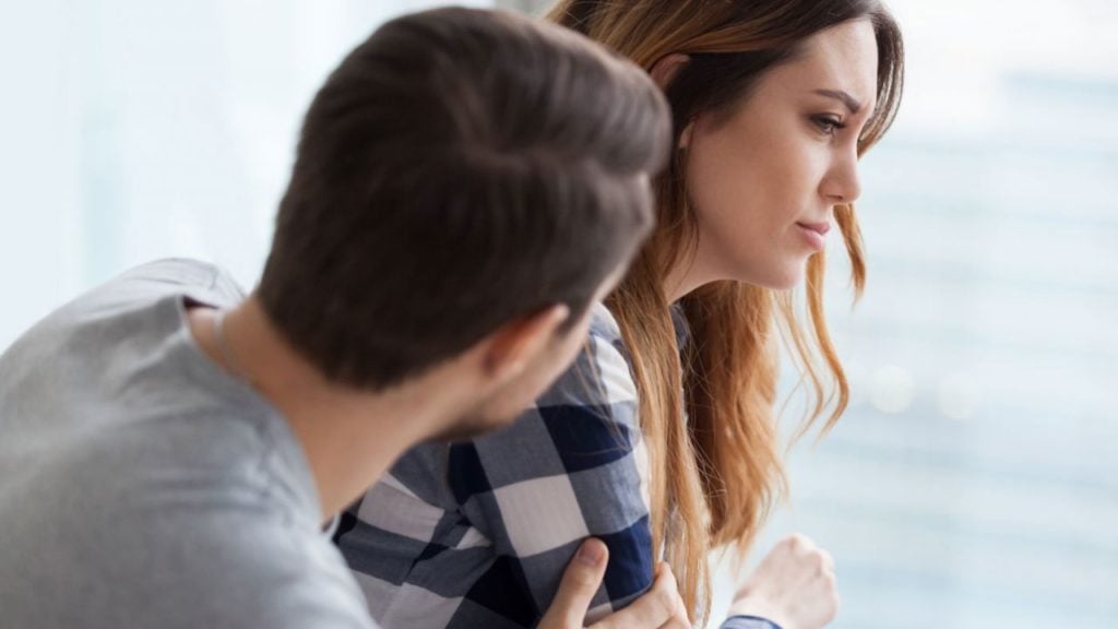 Foto ilustrativa de um casal triste, em que o homem está voltado à mulher, sentados com expressão triste em um fundo acinzentado