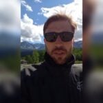 Fred Petla em vídeo gravado por câmera frontal de celular, com as montanhas do Alasca ao fundo da imagem, em dia de céu azul com nuvens brancas