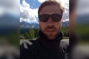 Fred Petla em vídeo gravado por câmera frontal de celular, com as montanhas do Alasca ao fundo da imagem, em dia de céu azul com nuvens brancas