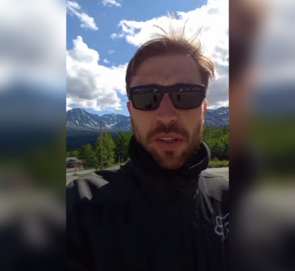 Fred Petla em vídeo gravado por câmera frontal de celular, com as montanhas do Alasca ao fundo da imagem, em dia de céu azul com nuvens brancas